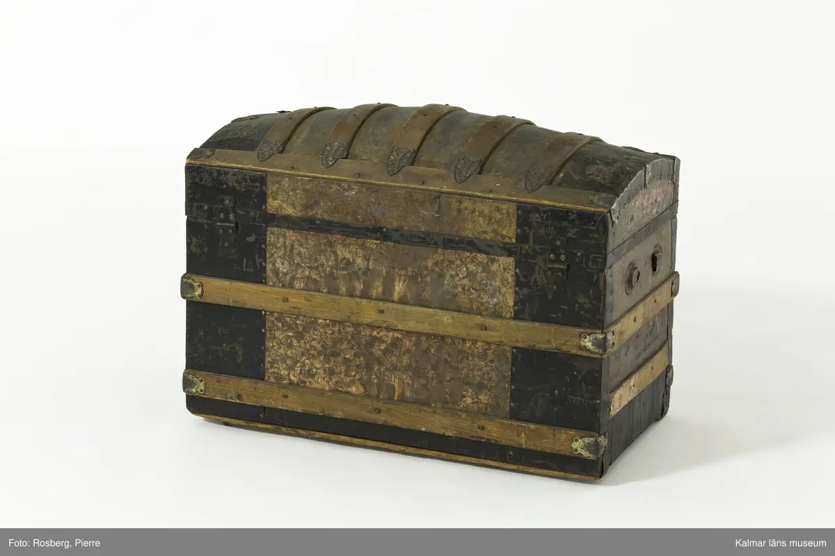 KLM 28210. Koffert, trä, metall, papp. Kista av trä med metallbeslag, välvt lock, lås, två spännen och beslag i form av sköldar med blommor. Delvis svartmålad med spår av gul färg. Handtag har suttit på gavlarna, båda saknas. Kofferten har haft fyra hjul av troligtvis mässing på underredet, endast ett av dessa är kvar. Invändigt klädd med papp med småblommigt mönster i grönt på gul botten. På lockets insida två fack. det högra facket har ett papper klistrat i botten med mönster i orange, blått och gult på grön botten och blommig bård i blått och grönt. På höger gavel påskrift: ? LSTRÖM ? SWEDEN.