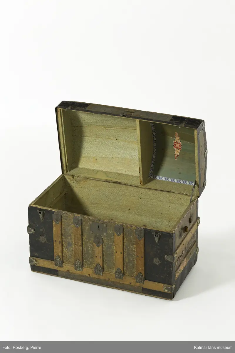KLM 28210. Koffert, trä, metall, papp. Kista av trä med metallbeslag, välvt lock, lås, två spännen och beslag i form av sköldar med blommor. Delvis svartmålad med spår av gul färg. Handtag har suttit på gavlarna, båda saknas. Kofferten har haft fyra hjul av troligtvis mässing på underredet, endast ett av dessa är kvar. Invändigt klädd med papp med småblommigt mönster i grönt på gul botten. På lockets insida två fack. det högra facket har ett papper klistrat i botten med mönster i orange, blått och gult på grön botten och blommig bård i blått och grönt. På höger gavel påskrift: ? LSTRÖM ? SWEDEN.