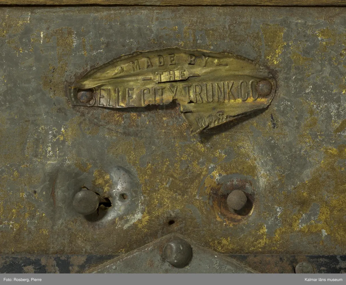 KLM 28210. Koffert, trä, metall, papp. Kista av trä med metallbeslag, välvt lock, lås, två spännen och beslag i form av sköldar med blommor. Delvis svartmålad med spår av gul färg. Handtag har suttit på gavlarna, båda saknas. Kofferten har haft fyra hjul av troligtvis mässing på underredet, endast ett av dessa är kvar. Invändigt klädd med papp med småblommigt mönster i grönt på gul botten. På lockets insida två fack. det högra facket har ett papper klistrat i botten med mönster i orange, blått och gult på grön botten och blommig bård i blått och grönt. På höger gavel påskrift: ? LSTRÖM ? SWEDEN.