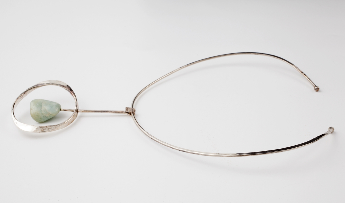 Halsklave bestående av en tynn sølvstreng, rund foran men etterhvert uthamret for å passe anatomisk rundt halsen. Klaven går ikke sammen bak og ender i to kuler. Fra klaven henger et anheng som består av en tynn stang med en lysegrønn sten, amazonitt. Til stangen er det også festet en hamret ring som omgir stenen.