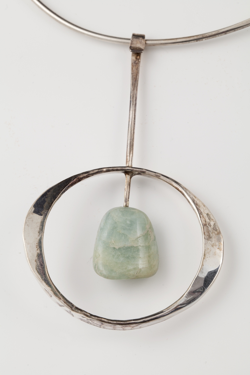 Halsklave bestående av en tynn sølvstreng, rund foran men etterhvert uthamret for å passe anatomisk rundt halsen. Klaven går ikke sammen bak og ender i to kuler. Fra klaven henger et anheng som består av en tynn stang med en lysegrønn sten, amazonitt. Til stangen er det også festet en hamret ring som omgir stenen.