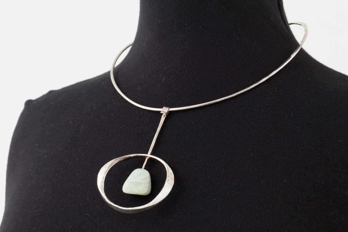 Halsklave bestående av en tynn sølvstreng, rund foran men etterhvert uthamret for å passe anatomisk rundt halsen. Klaven går ikke sammen bak og ender i to kuler. Fra klaven henger et anheng som består av en tynn stang med en lysegrønn sten, amazonitt. Til stangen er det også festet en hamret ring som omgir stenen.