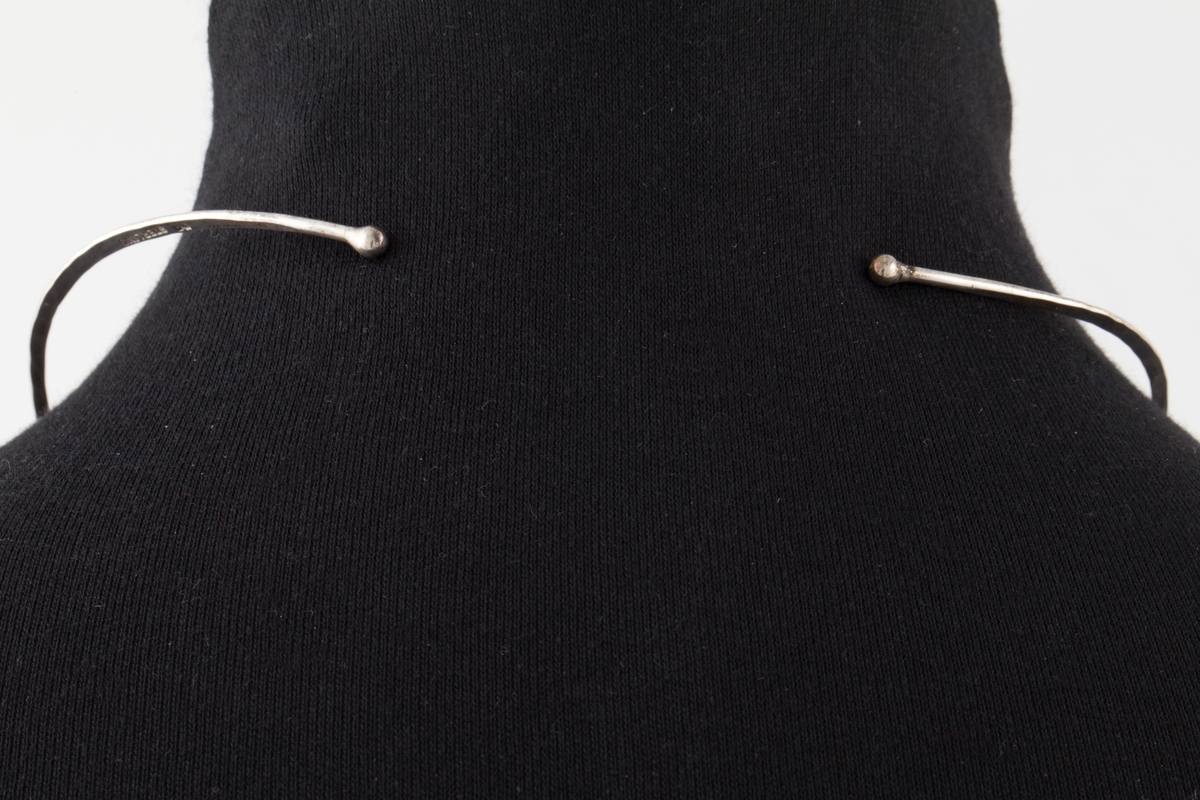 Halsklave bestående av en tynn sølvstreng, rund foran men etterhvert uthamret for å passe anatomisk rundt halsen. Klaven går ikke sammen bak og ender i to kuler. Fra klaven henger et anheng som består av en tynn stang med en lysegrønn sten, amazonitt. Til stangen er det også festet en hamret ring som omgir stenen.