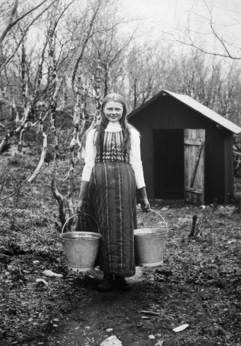 "Kvenpike". En ung kven jente står på stien med to bøtter full av vann i hendene. Brønnen (brønnhuset) ses i bakgrunnen. Rund den unge kvinnen vækser bjørketrær, det er tidlig på sommer og trærne holder på og sprette. Jenten er kledd i kjole med stripete vest og lys bommulkjorte.