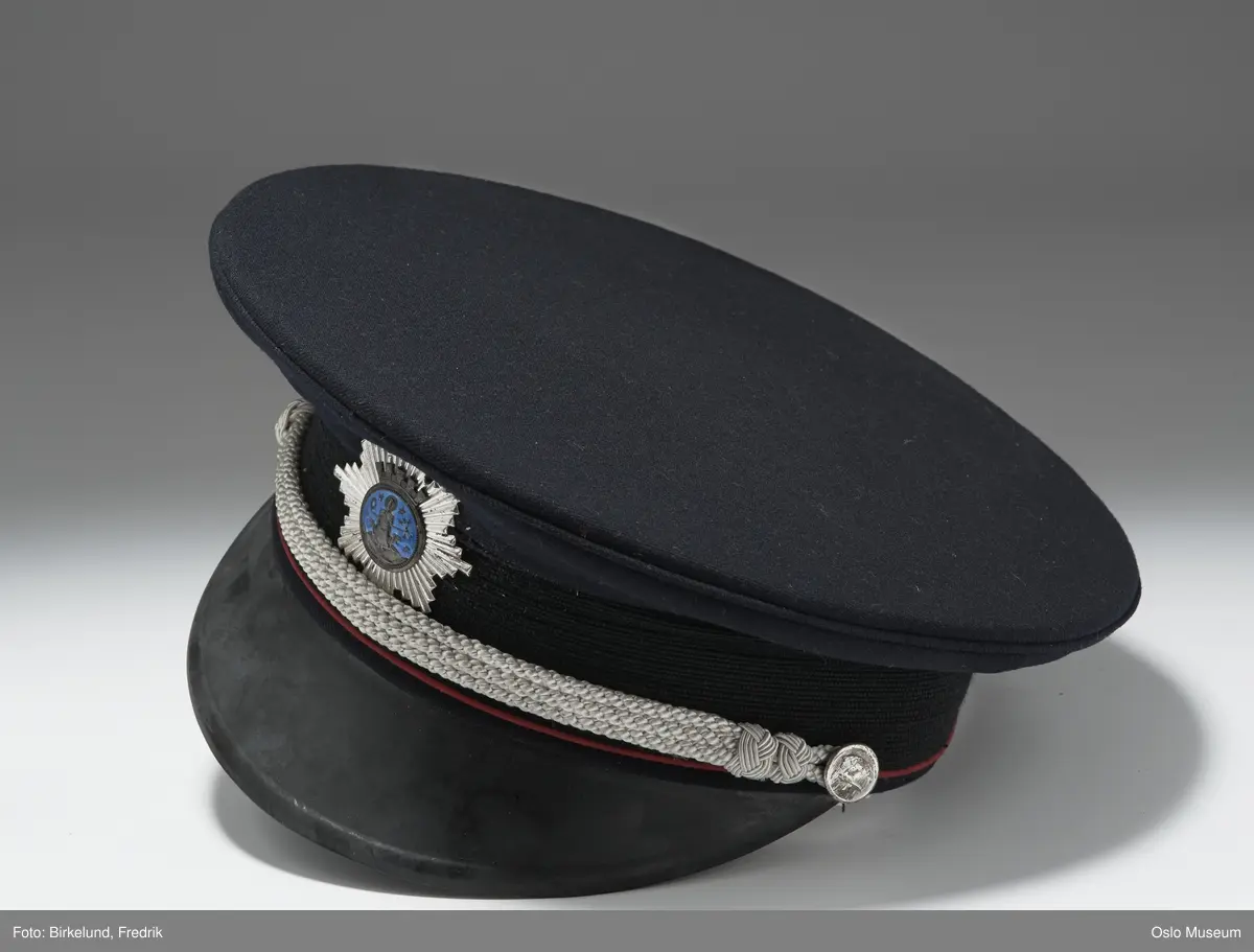 A.Jakke av mørkeblått ullstoff:Ombrettet krave m.lapeller,lange armer,lukning foran m. fire forgylte messingknapper m. brann- vesenets merke:brannhjelm m. hakke.To brystlommer m. fold og klaff m. knapp,to stikklommer m. klaff,fra brystlommene til nedkanten innsnitt.Skulderklaffer m. sølvtresser,kantet m. røde ullbånd,knapp og tre forsølvede stjerner.RESTEN,SE HÅNDARKIV. Forts.21:L=109,5cm  C.Uniformslue:H=13,5cm  D og E som C. F.Skyggelue:H=9cm LxB=26,5x25,7cm