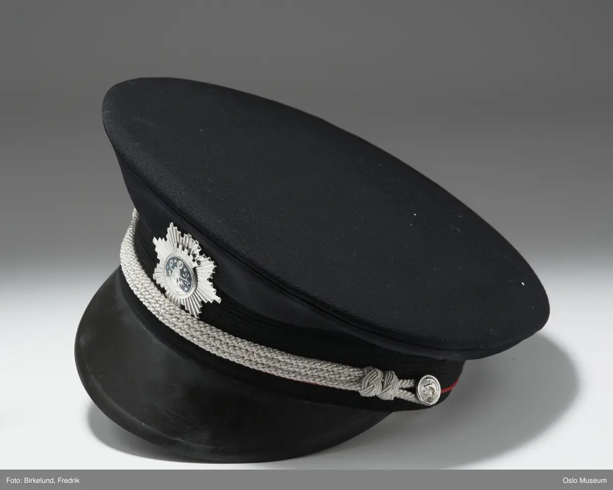 A.Jakke av mørkeblått ullstoff:Ombrettet krave m.lapeller,lange armer,lukning foran m. fire forgylte messingknapper m. brann- vesenets merke:brannhjelm m. hakke.To brystlommer m. fold og klaff m. knapp,to stikklommer m. klaff,fra brystlommene til nedkanten innsnitt.Skulderklaffer m. sølvtresser,kantet m. røde ullbånd,knapp og tre forsølvede stjerner.RESTEN,SE HÅNDARKIV. Forts.21:L=109,5cm  C.Uniformslue:H=13,5cm  D og E som C. F.Skyggelue:H=9cm LxB=26,5x25,7cm