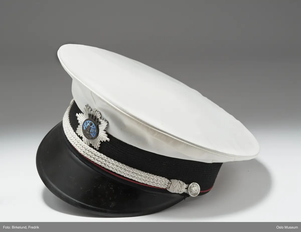 A.Jakke av mørkeblått ullstoff:Ombrettet krave m.lapeller,lange armer,lukning foran m. fire forgylte messingknapper m. brann- vesenets merke:brannhjelm m. hakke.To brystlommer m. fold og klaff m. knapp,to stikklommer m. klaff,fra brystlommene til nedkanten innsnitt.Skulderklaffer m. sølvtresser,kantet m. røde ullbånd,knapp og tre forsølvede stjerner.RESTEN,SE HÅNDARKIV. Forts.21:L=109,5cm  C.Uniformslue:H=13,5cm  D og E som C. F.Skyggelue:H=9cm LxB=26,5x25,7cm