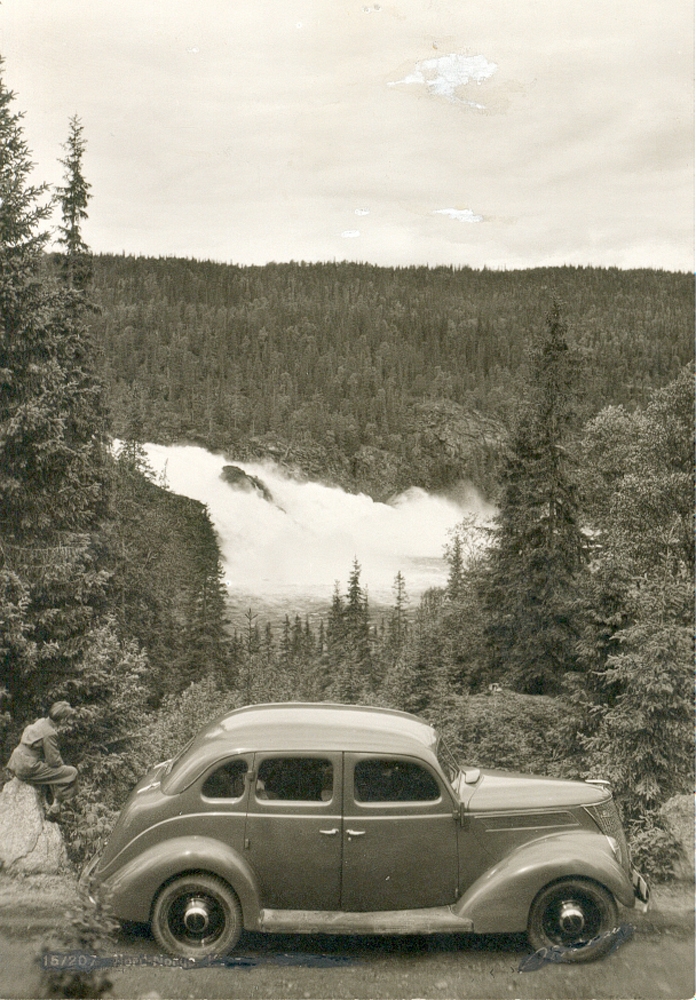 Postkort. Stabbforsen, Korgen. 1945. Mann sitter på en stein, bil i forgrunn. Normann Kunstforlag.