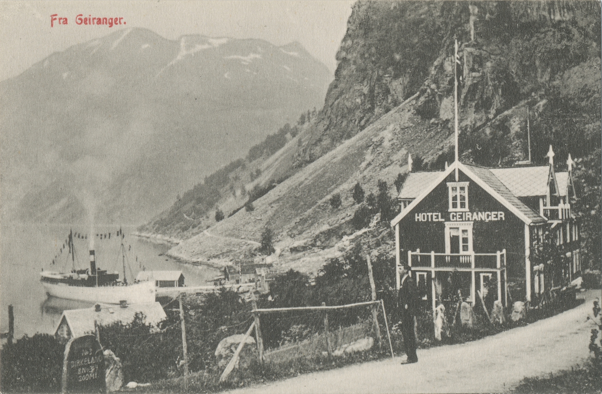 Prospektkort av hotell Geiranger med Geirangerfjorden og et skip til kai i bakgrunnen. En mann står og ser på utsikten fra en vei foran hotellet.
