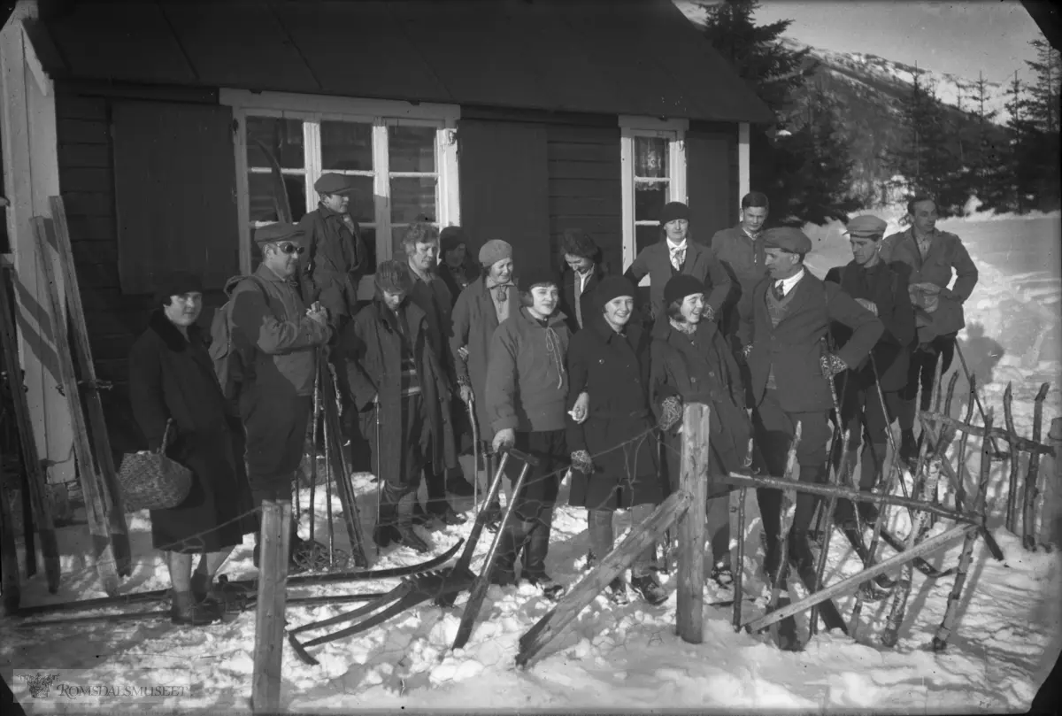 Personar med ski og sparkstøtting utan for ei hytte.