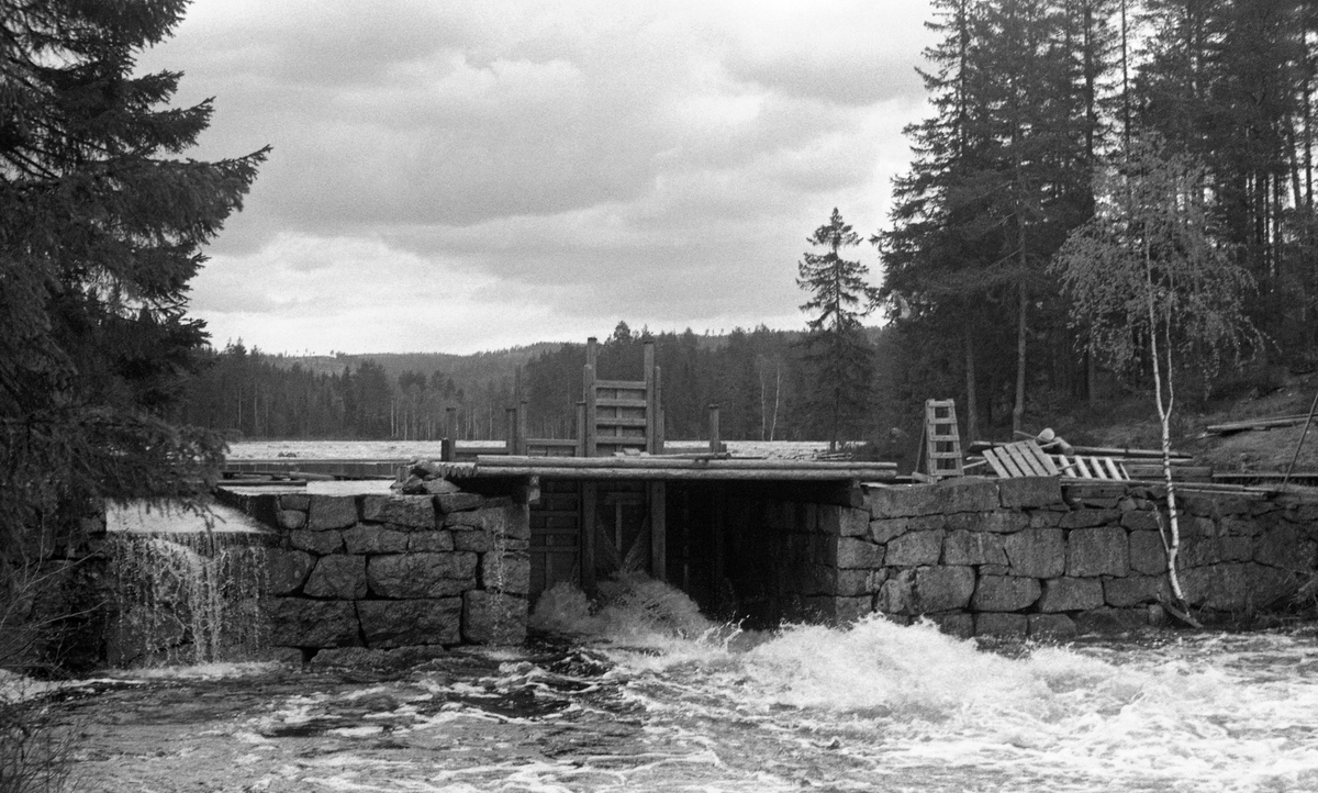 Fjæstaddammen i Malungsvassdraget i Romedal allmenning i Hedmark, fotografert i motstrøms retning våren 1960. Dette var en murt steindam, oppført der hvor Fjæstadsætertjennet hadde sitt utløp i ei cirka halvannen kilometer lang å som førte vannet ned mot Rasasjøen. Fjæstaddammen var en såkalt lukedam hvor det kunne bygges opp et betydelig reservoar av vann for fløtinga i den nedenforliggende åa ved å senke fire luker - trelemmer - mot verikaltstilte bjelker på motstrøms side av damåpningen. Lukene dannet en vegg som reduserte avrenninga til et minimum. På denne måten kunne fløterne etablere et vannreservoar som seinere kunne tappes, slik at vannføringa i åa ble høy nok til at tømmeret som var levert på Fjæstadtjennet kunne flyte noenlunde uhindret nedover til Rasasjøen. Da dette fotografiet ble tatt var dammen antakelig full. Ei av de fire lukene var litt hevet for å slippe ut overflødig vann, men dammen var ennå ikke åpnet for fløting.