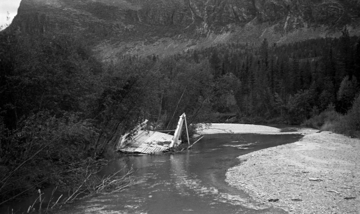 Flommen i slutten av mai 1958, som ble forsterket av kraftige regnbyger, gjorde store skader i Nord-Østerdalen. I Tylldalen vasket elva Tysla med seg fire-fem kilometer av bygdevegen og flomvannet som trengte inn på dyrket mark vasket bort gjødsel og la i stedet igjen mye grus. Elva rev også med seg brua ved kirka og flyttet på sagbruket ved Hokstad-gardene. Tylldal snekkerverksted ble også hardt rammet. I Tysladammen brast lensa, og cirka 3 000 kubikkmeter med tømmer forsvant utfor den nedenforliggende fossen uten at fløterne hadde kontroll med det som foregikk. Dette fotografiet er sannsynligvis tatt i den søndre delen av Tylldalen etter at flommen var over. Vi ser ei havrert bru som lå igjen i elveløpet, der det også var store grusører. Langs elvebreddene var det tydelig at lauvskogen hadde vært utsatt for hurtigstrømmende vannmasser som hadde fått mange av trærne til å bikke.