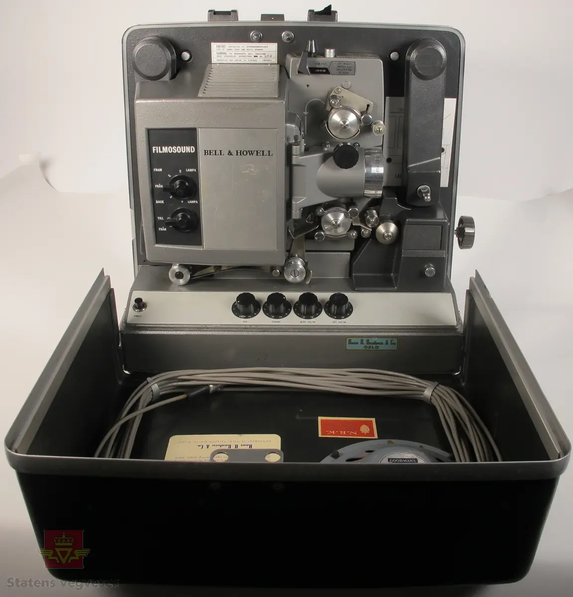BELL & HOWELL MODEL 642. 16 mm filmfremviser i mørk grå koffert av glassfiber, med defekt bærehåndtak. Den ene delen av kofferten har høyttaler av merket Goodmans, ledning for tilkobling av høyttaler og to klistremerker med påskrifter. Filmfreviseren har eget panel for justering av lyden. Bak på kofferten er det to skiltplater med innskrifter, MODEL 642 SK SERIAL No. K418 220V-1100 W - 50 ~ MADE IN ENGLAND og HANS H. KNUTSEN & CO. OSLO TYPE 642 No 418 P.VOLT 200/250 50 ~ S.VOLT AMP. 1200W. Veske, strømledning, ekstra lyspære og to tomme filmruller følger med filmfremviseren.