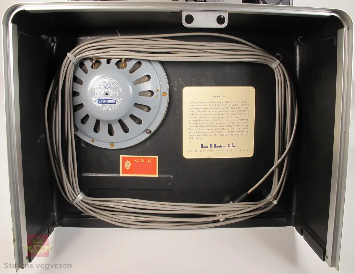 BELL & HOWELL MODEL 642. 16 mm filmfremviser i mørk grå koffert av glassfiber, med defekt bærehåndtak. Den ene delen av kofferten har høyttaler av merket Goodmans, ledning for tilkobling av høyttaler og to klistremerker med påskrifter. Filmfreviseren har eget panel for justering av lyden. Bak på kofferten er det to skiltplater med innskrifter, MODEL 642 SK SERIAL No. K418 220V-1100 W - 50 ~ MADE IN ENGLAND og HANS H. KNUTSEN & CO. OSLO TYPE 642 No 418 P.VOLT 200/250 50 ~ S.VOLT AMP. 1200W. Veske, strømledning, ekstra lyspære og to tomme filmruller følger med filmfremviseren.