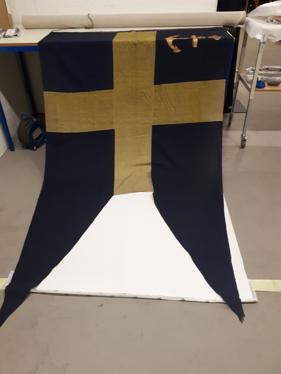 Lotsflagga av bomull. Svensk tvåtungad flagga med Kgl. Lotsverkets emblem med krona över i inre fältet. Tamp med lekare.