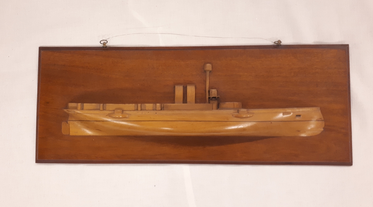 Fartygsmodell med roder, mast, 2 fyrkantiga skorstenar och  brygga. Fästad til väggplan.