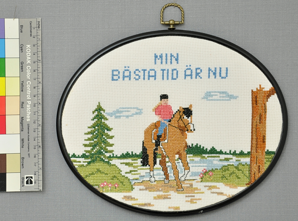 Oval tavla med svart plastram. Broderi i DMC-garn på blekt aidaväv. Korsstygn och efterstygn i svart och olika bruna, rosa, blå och gröna nyanser.

Motiv: Text "Min bästa tid är nu". Flicka på häst på skogsväg. Gran och trädstam; granskog och vattendrag i bakgrunden.

