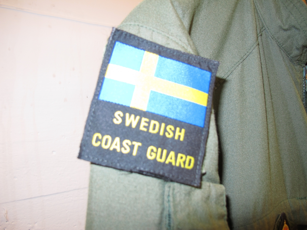 Overall, grön med emblém. På höger ärm svensk flagga med texten "Swedish coast guard" undertill. 
På höger ärm Kustbevakningens örnhuvud och gradbeteckning.
På höger bröst emblém: Kustbevakningen flyg.
På vänter bröst rund EU-flagga och där under textband "Kustbevakningen". Av bomullsmix.