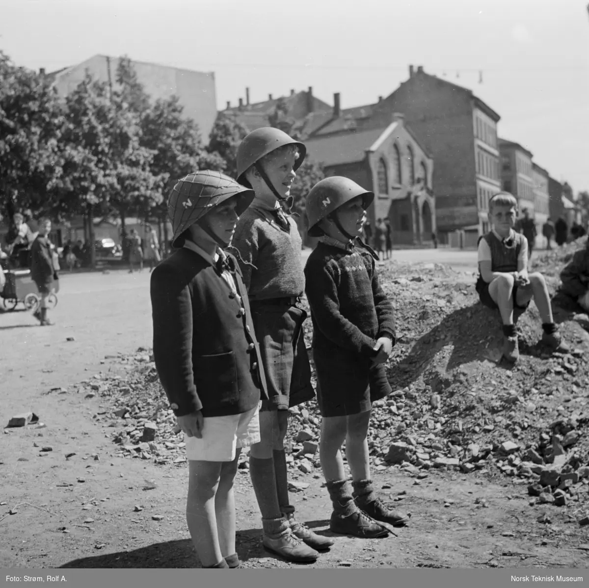 Tre små gutter med soldathjelmer, kortbukser og ellers i finklær, stående ved haug med stein og tegl, Oslo, juni 1945.