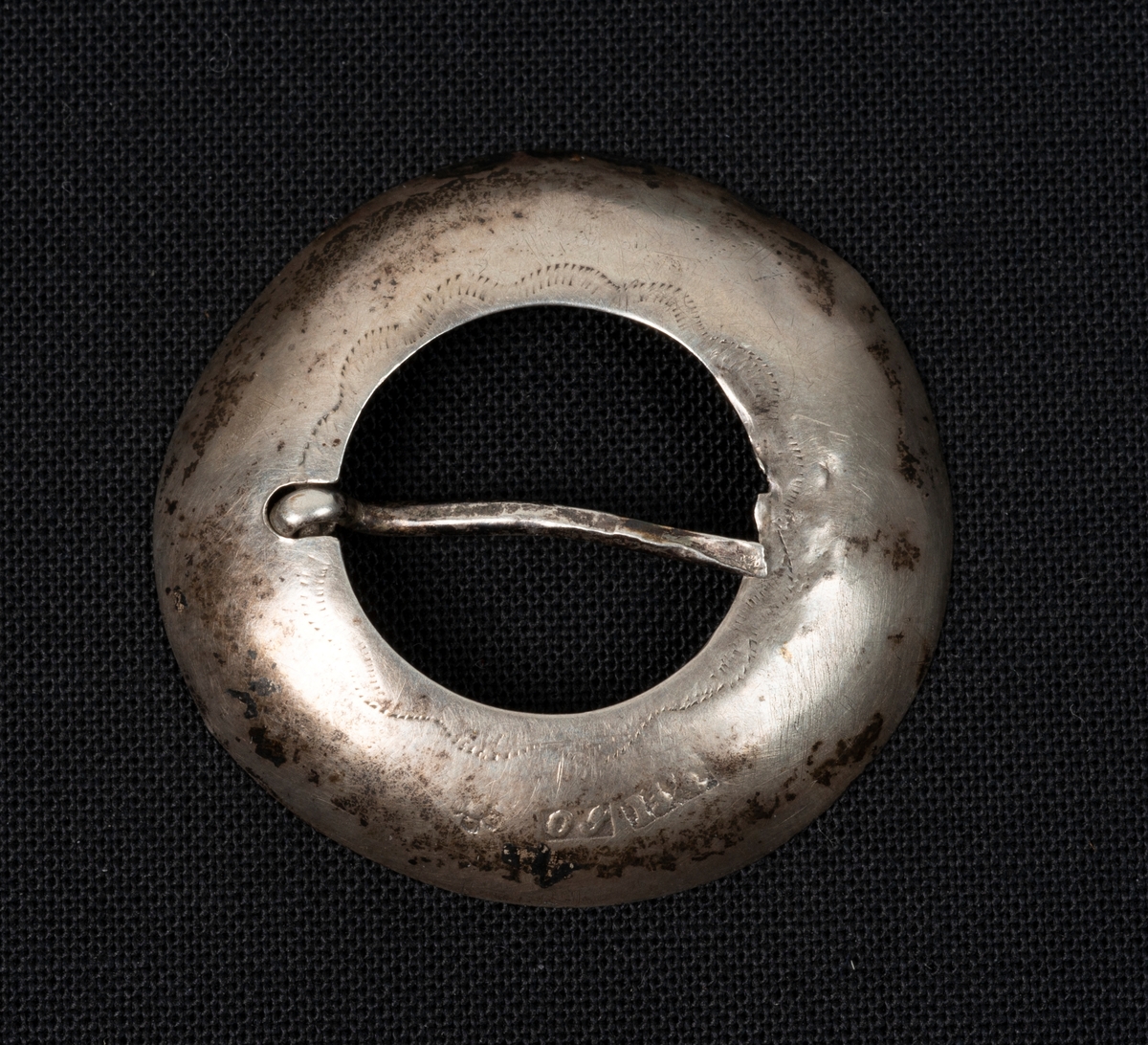 Dette er ein flat ring av sølv, laga av ei sølvplate. Den er litt gravert og har fått ei litt bua form. Ingen lauv. Festes med ei tann gjennom stoffet. 