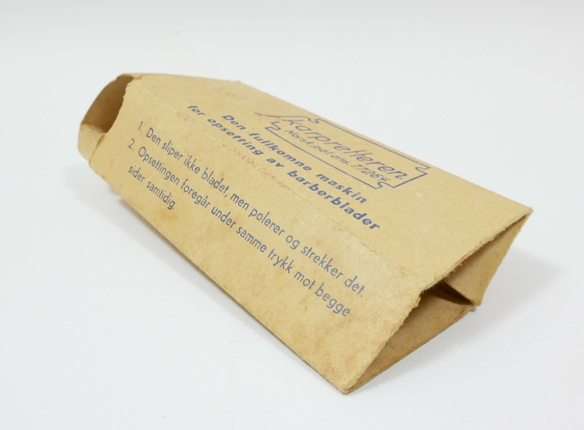 Rektangulær kartongemballasje med påskrift i blått. 

Påskrift:
Skarpretteren // Norsk pat.ans 77264  // Den fullkomne maskin // for opsetting av barberblader 

Påskrift: 
1. Den sliper ikke bladet, men oplerer og strekker det. 
2. påsettingen foregår under samme trykk mot begge // sider samtidig 

Påskrift: 
BRUKSANVISNING: // a. Stryk en gang imellem litt fett, olje eller såpe på slipelegeme. // b. Legg bladet på plass og lukk apparatet. // c. Trykk meget lett på lokket og beveg tappen frem og tilbake // 20 til 25 ganger. - Prøv så bladet og gjenta operasjonen, om // nødvendig, inntil eggen er skarp // Med litt erfaring opnår man et utmerket resultat. 

Påskrift: 
3. Ved riktig behandling forebygges at bladet blir // så tynt i eggen at denne bøier sig og danner // den såkalte rå- egg
