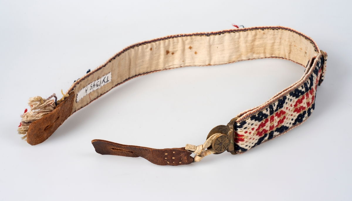 Buksehengsler av vevde bånd. Hengslene er fôra med tekstil og festene er lagd av skinn og metall. Festene i den ene enden er skjult av en frynsedusk.