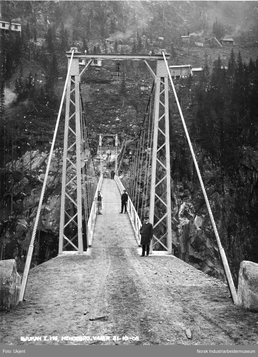 Hengebroen mellom Vaaer og Vemork er ferdig. Fem menn stående på boen, sannsynligvis ingeniører. Brakker på Vemorksiden ligger i fjellsiden.