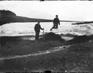 Två officerare från kanonbåten HMS Svensksund på jakt på Spetsbergen. En man med gevär, sannolikt löjtnant Lagercrantz, står nedanför en låg glaciärkant och tittar mot fotografen. På glaciären står en man på knä med renhorn i handen och tittar mot höger. Han är kapten Hamilton.