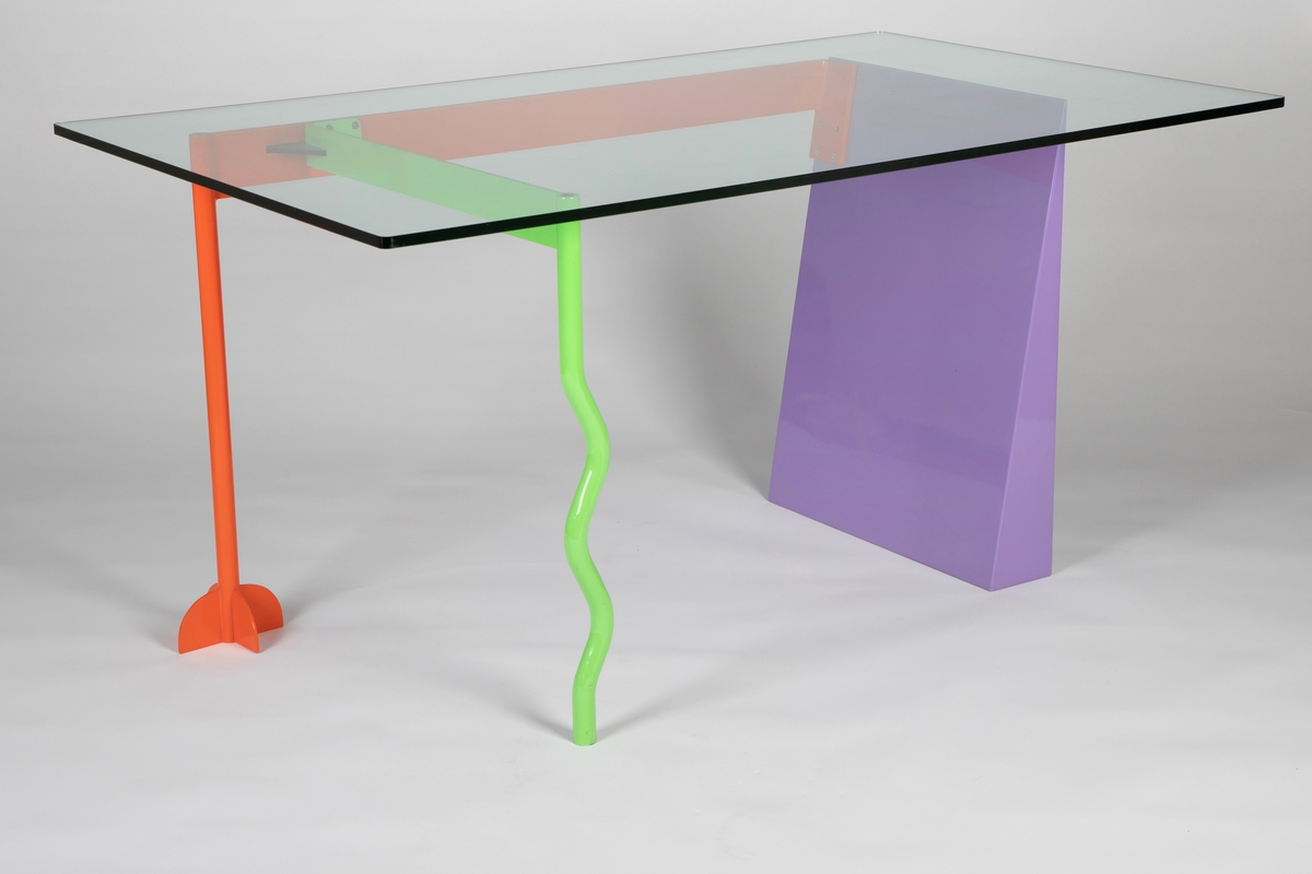 Bordplate i tykt, klart glass på et inntrukket understell av lakkert metall og plastlaminert tre. Foran til venstre et tynt, lysgrønt bølgete bein med en øvre tverrligger festet med et svart element til et oransje bakbein med brudt fotsokkel. Det oransje beinet er del av en lengre oransje tverrligger i skrivebordets bakre breddekant. Høyre side av bordet er støttet på en lyslilla plate som på sin ytre side står 90 grader fra underlaget, mens den skrår oppover fra underlaget på sin indre side; den er altså bredere nederst enn øverst, der den er spiss, og har dermed rettvinklede trekanter i profil.