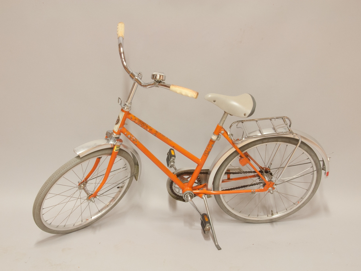 Barncykel, DBS modell "Flowers", orangefärgad med påklistrad blomdekor. Storlek 20". Fälgar märkta "Alumaxium", skärmar märkta "Elox alum.". Kromat styre och pakethållare, viss korrosion. 
Handtagen av PVC samt grå gummidäck, åldrade. Blocklås "Basta", avsågat.