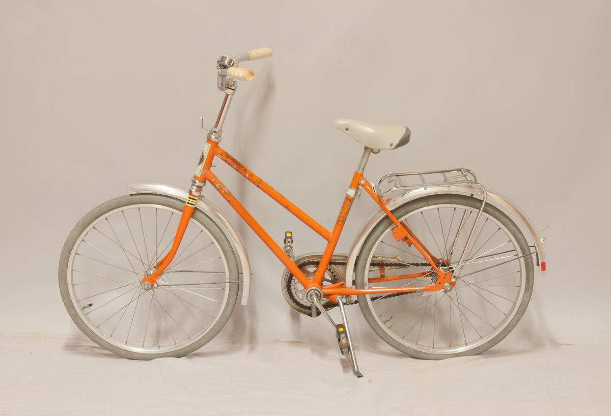Barncykel, DBS modell "Flowers", orangefärgad med påklistrad blomdekor. Storlek 20". Fälgar märkta "Alumaxium", skärmar märkta "Elox alum.". Kromat styre och pakethållare, viss korrosion. 
Handtagen av PVC samt grå gummidäck, åldrade. Blocklås "Basta", avsågat.