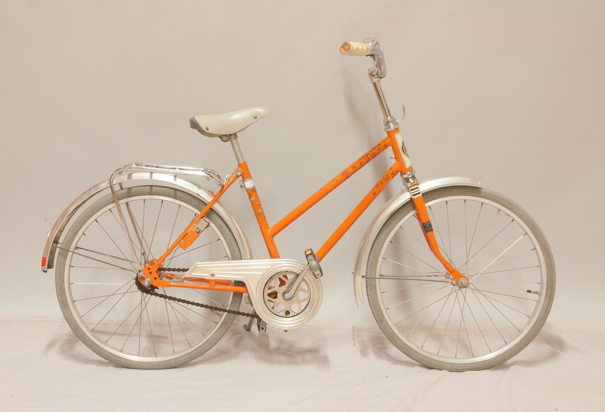 Barncykel, DBS modell "Flowers", orangefärgad med påklistrad blomdekor. Storlek 20". Fälgar märkta "Alumaxium", skärmar märkta "Elox alum.". Kromat styre och pakethållare, viss korrosion. 
Handtagen av PVC samt grå gummidäck, åldrade. Blocklås "Basta", avsågat.