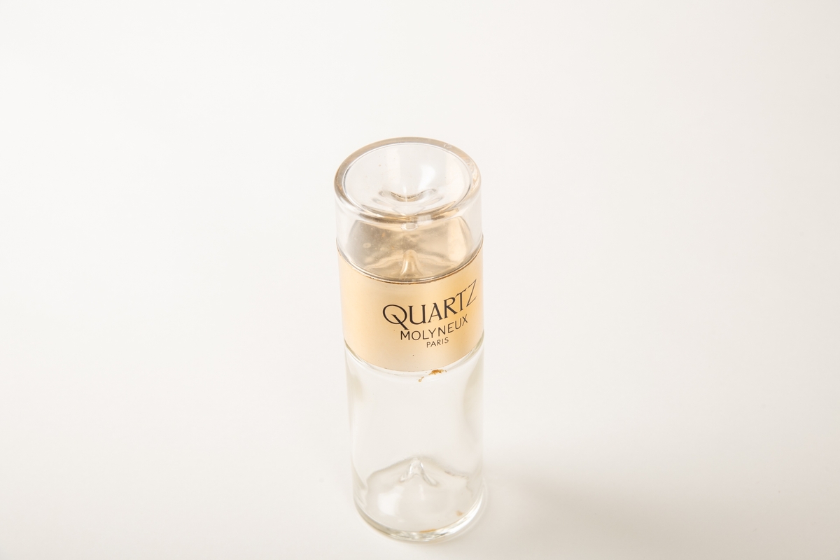 Sylindrisk flaske av tykt glass, med en oppstående dråpeform i bunnen og innv. i lokket.  Lokket er øvre del av sylinderen og har tekst:   EAU DE PARFUM 850 og QUARTZ MOLYNEUX Paris