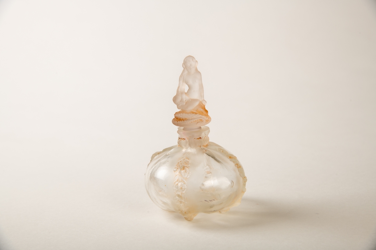 Rund krukke med fire blomsterranker i relieff ned siden. Liten munning med propp, hvorpå en  sittende kvinne på en slange. Inngravert:  Eva.