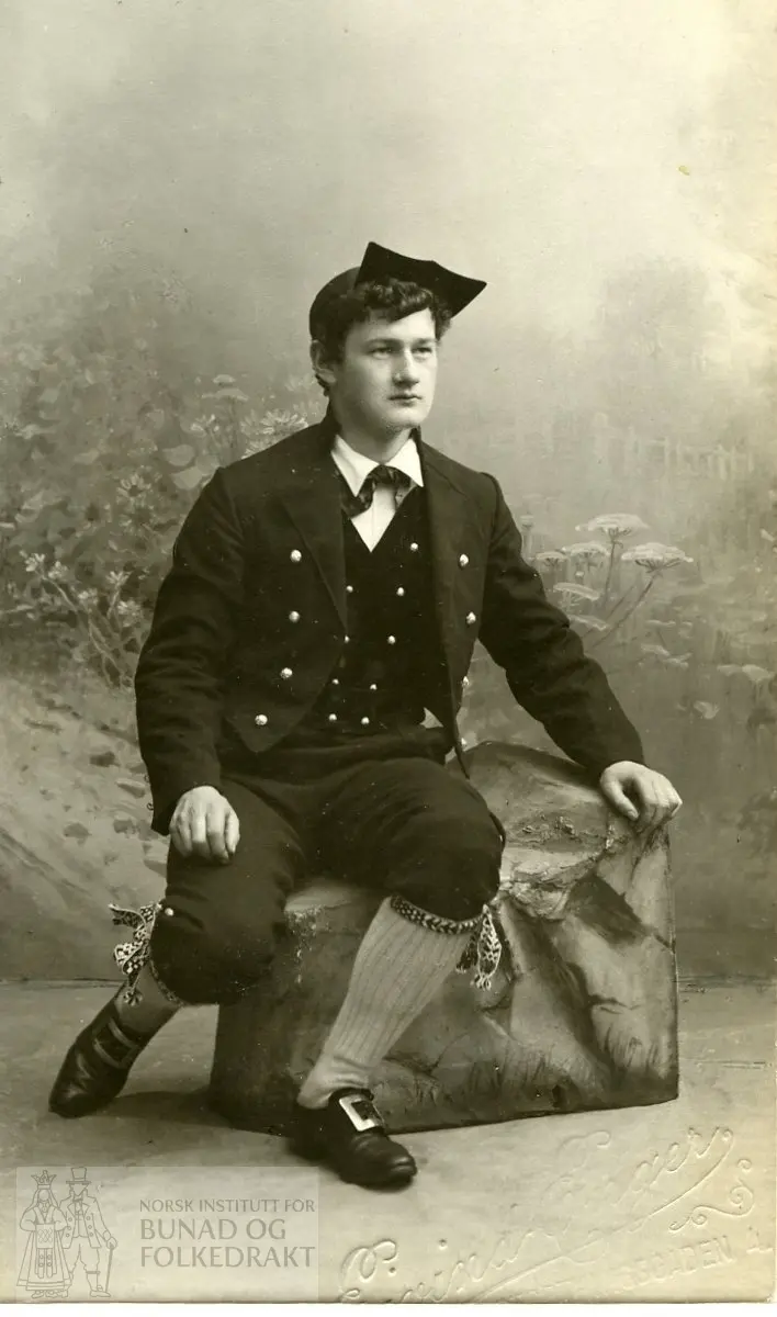 Mann kledd i stuttrøye og skjoldhuve frå Storelvdal. Han har silketørkle i halsen, dobbeltspent attknept vest, knebukse, kvite strikka strømper og vove sokkeband. Bilete teke hjå fotograf.