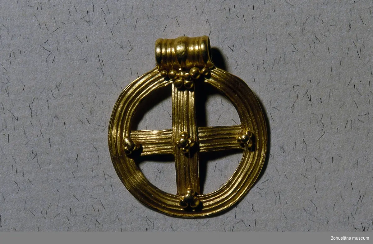 Ett hängsmycke, i orm av ett ringkors av guld med fast ögla. Korset är likarmat och placerat i en ring av samma bredd som korsarmarna. På framsidan är ytan dekorerad med längsgående, något oregelbunden räffling. Korsmitten och korsarmarnas anslutningspunkter dekorerade med granulering (små kulor). 
Smycket dateras till folkvandringstid, 400 - 600 e. Kr. och brukar kallas för "Rörakorset". 

Landskap BOHUSLÄN
LÖSFYND