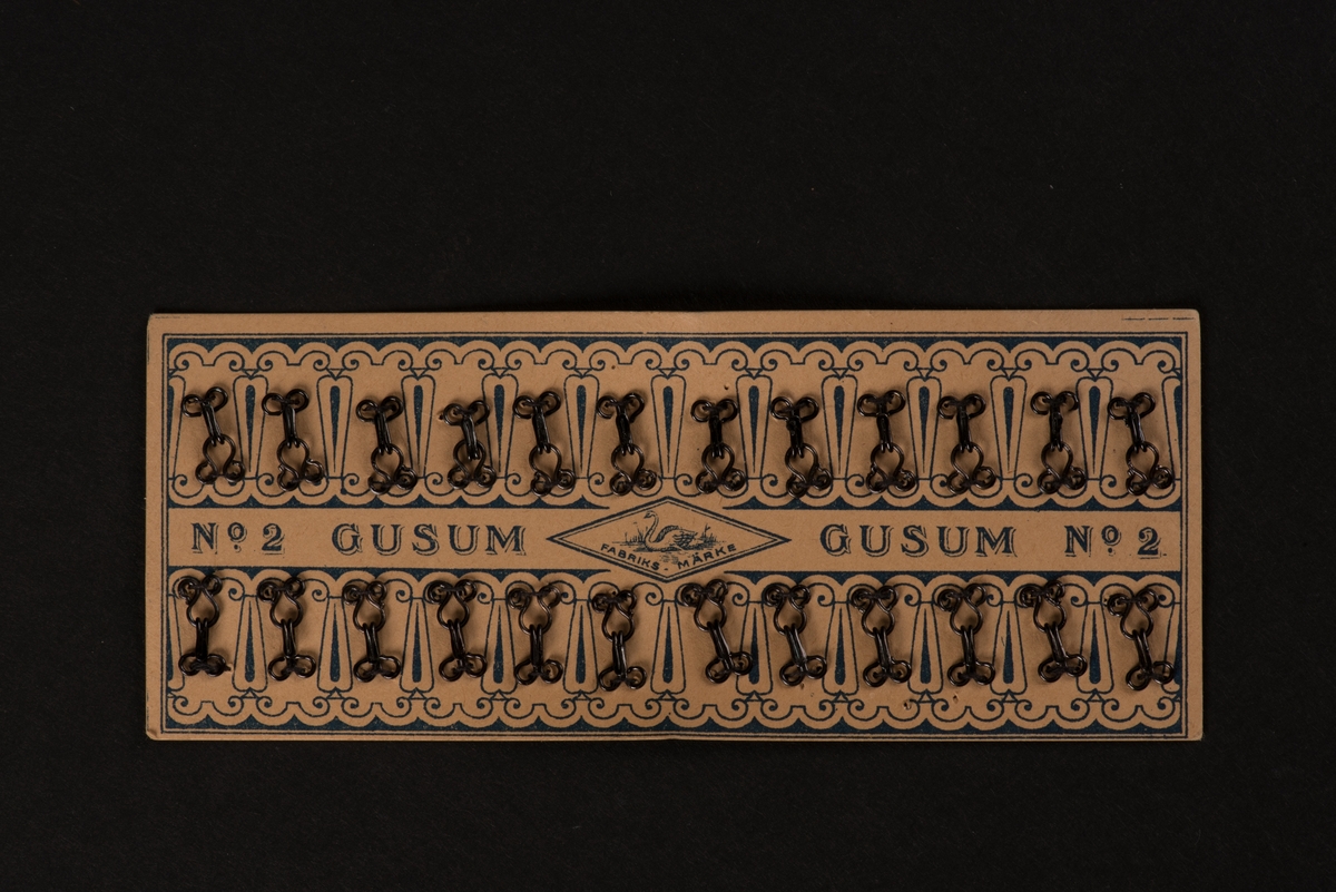 En karta av papp med fastsydda hyskor och hakar av metall. Två rader, med tjugofyra hakar och hyskor. Kartan är dekorerad med ett tryckt mönster, samt "GUSUM No 2". I mitten en svan över texten "FABRIKS-MÄRKE" inom en romb.