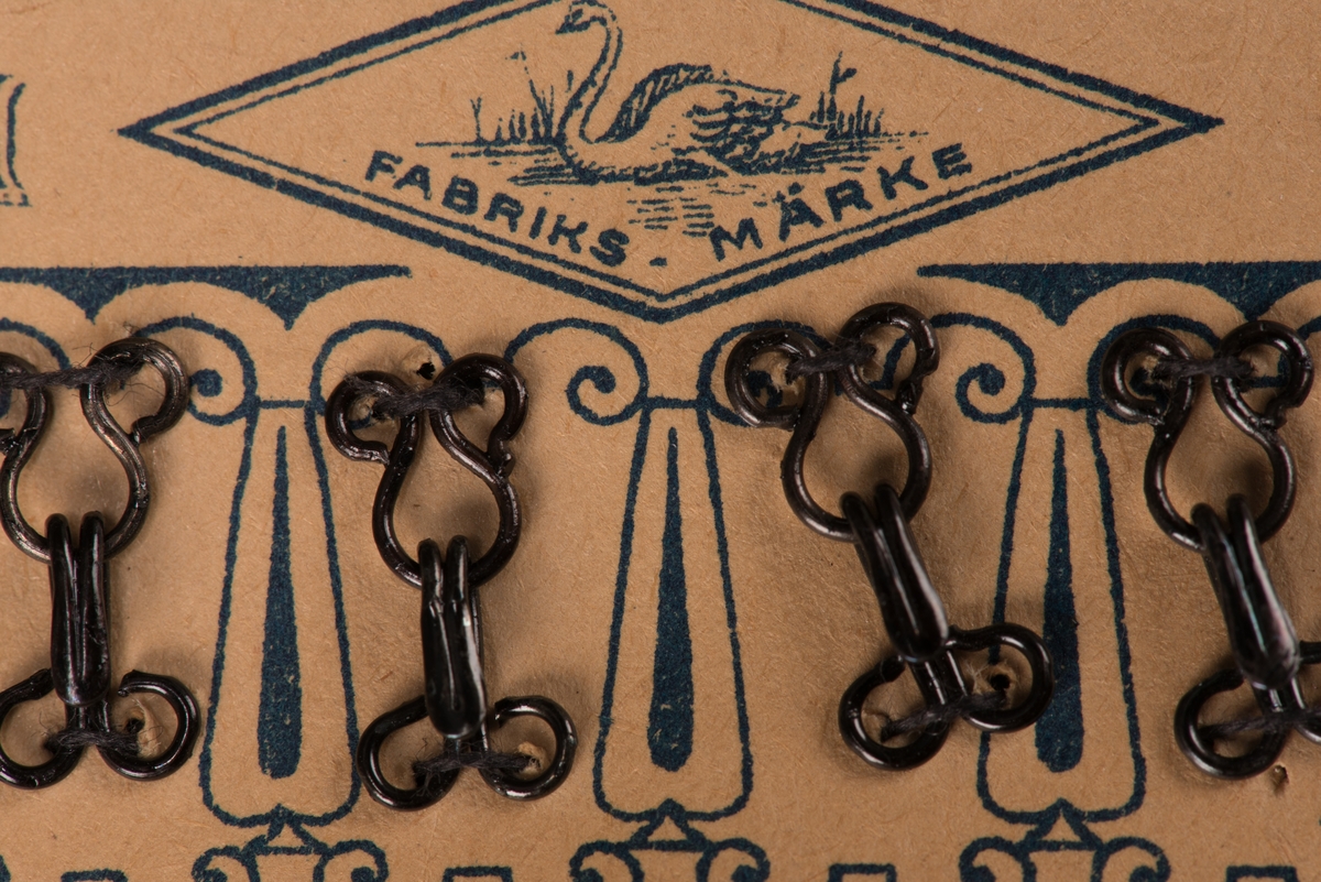 En karta av papp med fastsydda hyskor och hakar av metall. Två rader, med tjugofyra hakar och hyskor. Kartan är dekorerad med ett tryckt mönster, samt "GUSUM No 2". I mitten en svan över texten "FABRIKS-MÄRKE" inom en romb.