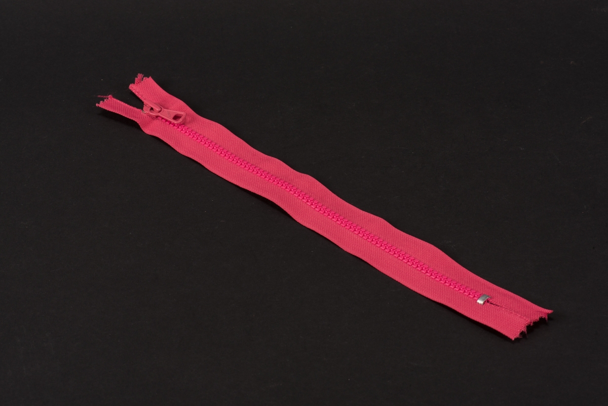 Ceriserosa dragkedja med textilband, häktor av plast och löpare och dragtapp av metall. På dragtappen inpräglat GUSUM.