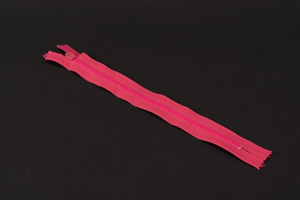 Ceriserosa dragkedja med textilband, häktor av plast och löpare och dragtapp av metall. På dragtappen inpräglat GUSUM.
