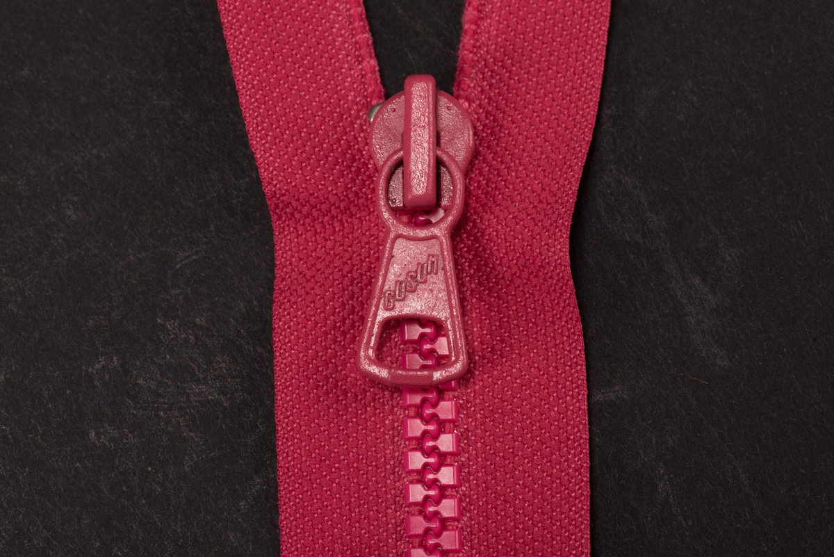Ceriserosa dragkedja med textilband, häktor av plast och löpare och dragtapp av metall. På dragtappen inpräglat GUSUM.