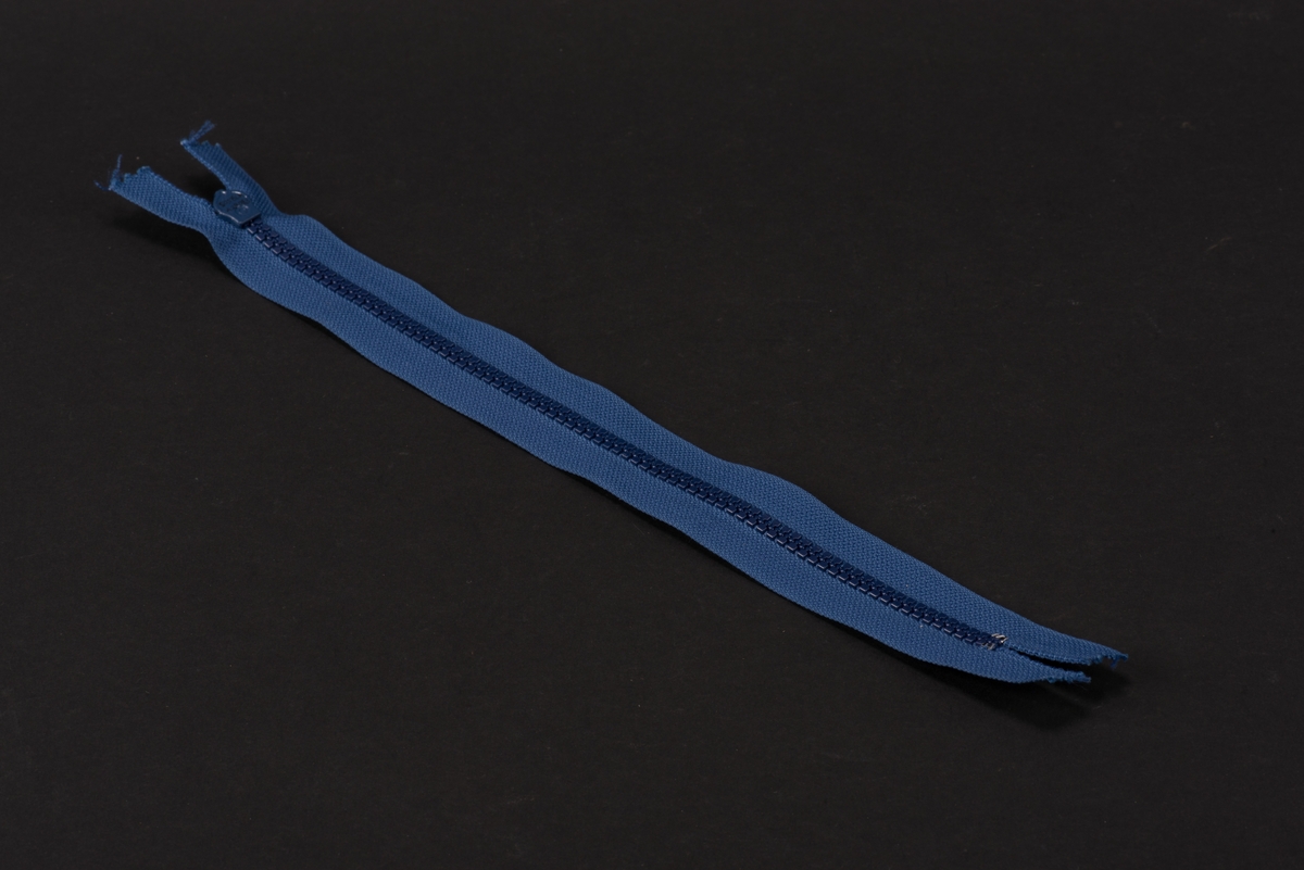 Blå dragkedja med textilband, häktor av plast och löpare och dragtapp av metall. På dragtappen inpräglat GUSUM.