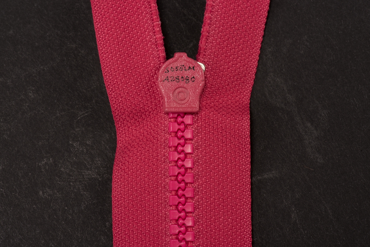 Ceriserosa dragkedja med textilband, häktor av plast och löpare och dragtapp av metall. På dragtappen inpräglat GUSUM.