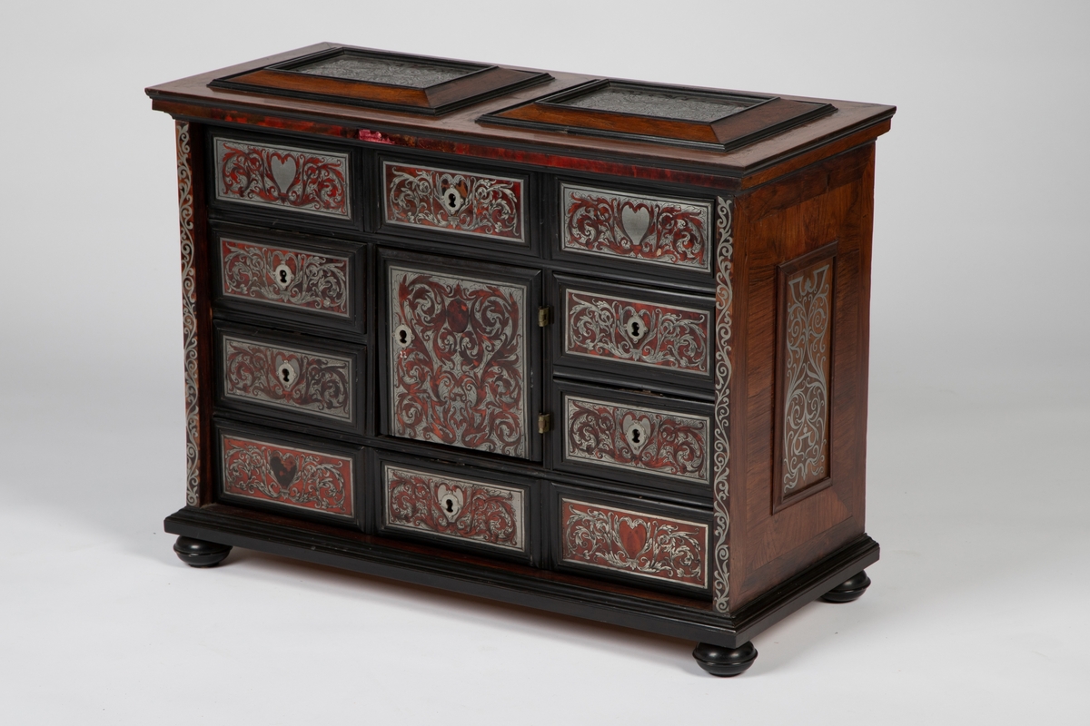 Kabinett med kuleben og profilert fotlist. Fronten har 10 skuffer omkring en dør i midten, innrammet av profilerte lister av ibenholt. Alle fyllinger har marketeri i "Boulle-teknikk", bestående av skilpadde og tinn. som er gravert med akantusranker. Sidelister, topplatens to fyllinger og sidenes to fyllinger har alle marketeri i tilsvarende teknikk med ranker. Profilert topplist med kant av skilpadde. Tre skuffer bak midtdøren.