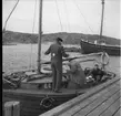 En träbåt med mast och inombordare ligger vid en brygga, troligen på väg att lägga ut. Fyra män ses ombord.