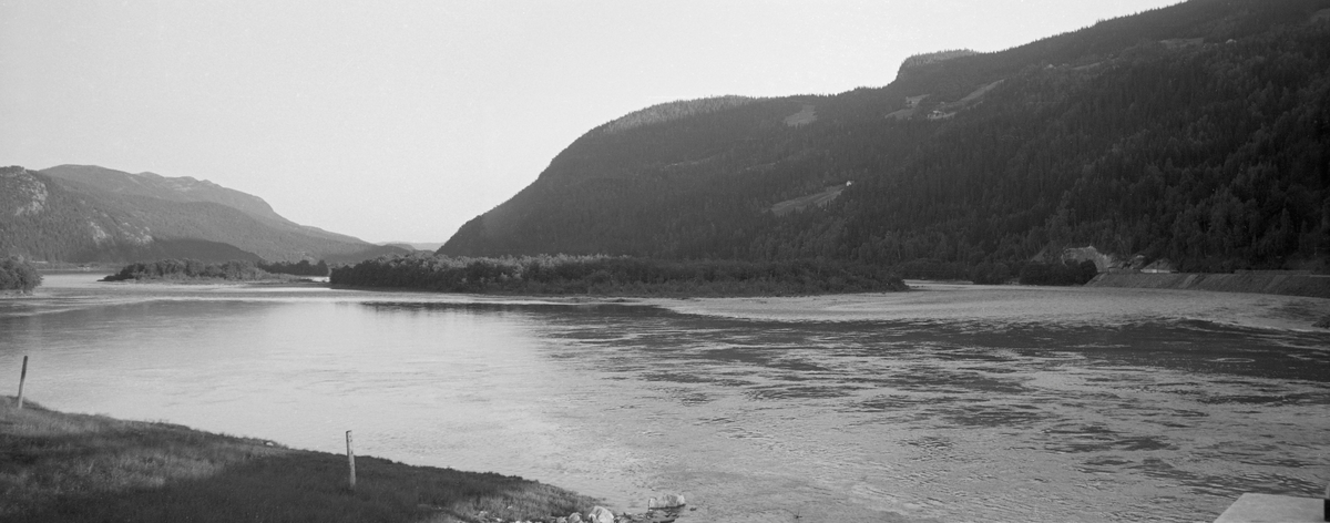 Elvelandskap i Ringebu i Gudbrandsdalen. Fotografiet ble tatt i medstrøms retning fra en posisjon ved Rankleivbruas østre brukar, altså sørover. Her renner Lågen forholdsvis stilleflytende rundt Gåsøya (nærmest) og Olstadøya. Mens elva her er vid, stilleflytende og fyller mesteparten av den flate dalbotnen, er liene på begge sider bratte og skogbevokste der det var jordsmonn nok til at det kunne vokse trær. Fotografen, som var funksjonær i Glomma fellesfløtingsforening, var opptatt av landskapselementer som vanskeliggjorde framdriften av tømmeret. Her gjaldt det nok særlig sandbankene elva hadde lagt igjen, der tømmerstokkene hadde lett for å strande og filtre seg sammen når vannstanden var synkende.