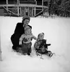 En kvinna med två barn leker i snön vid Rotundan i Jönköpings stadspark. Barnen är klädda i mönsterstickade tröjor och mössor med stor tofs.