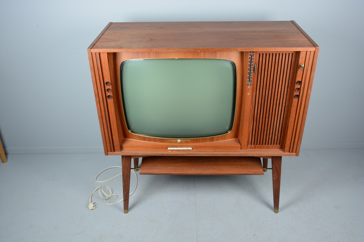TV/fjernsyn samt radio monterte i kabinett med skyvedører for skjuling av fjernsynet når det ikkje var i bruk. Beteningsknappar og elektrisk kabel. Kabinettet har liggande rektangulær form.