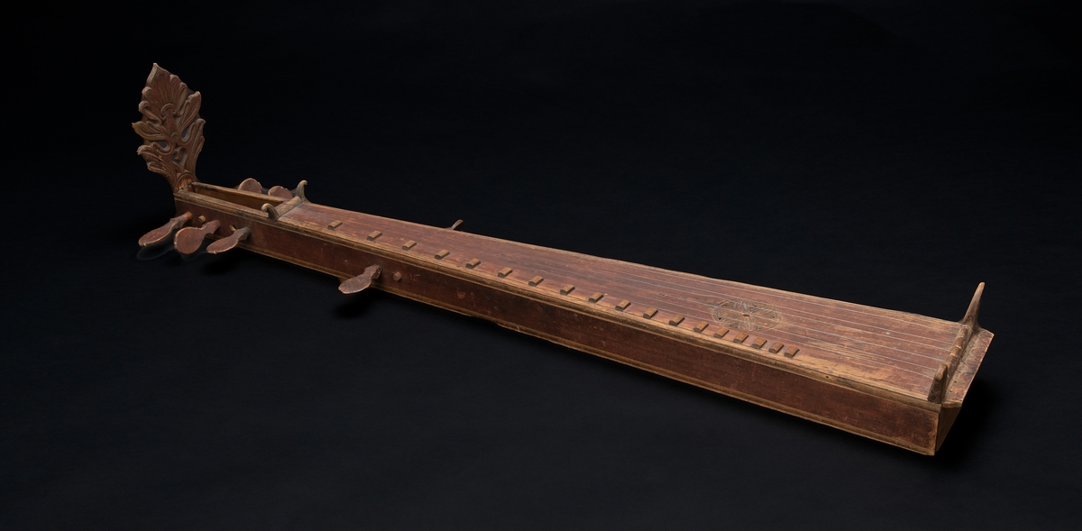 Langeleik av gudbrandsdalstype, med utskåret endestykke som har en inskripsjon på baksiden. Åtte strengeskruer av tre, hvorav den ene er brukket. Utskåret rose midt på kassen.