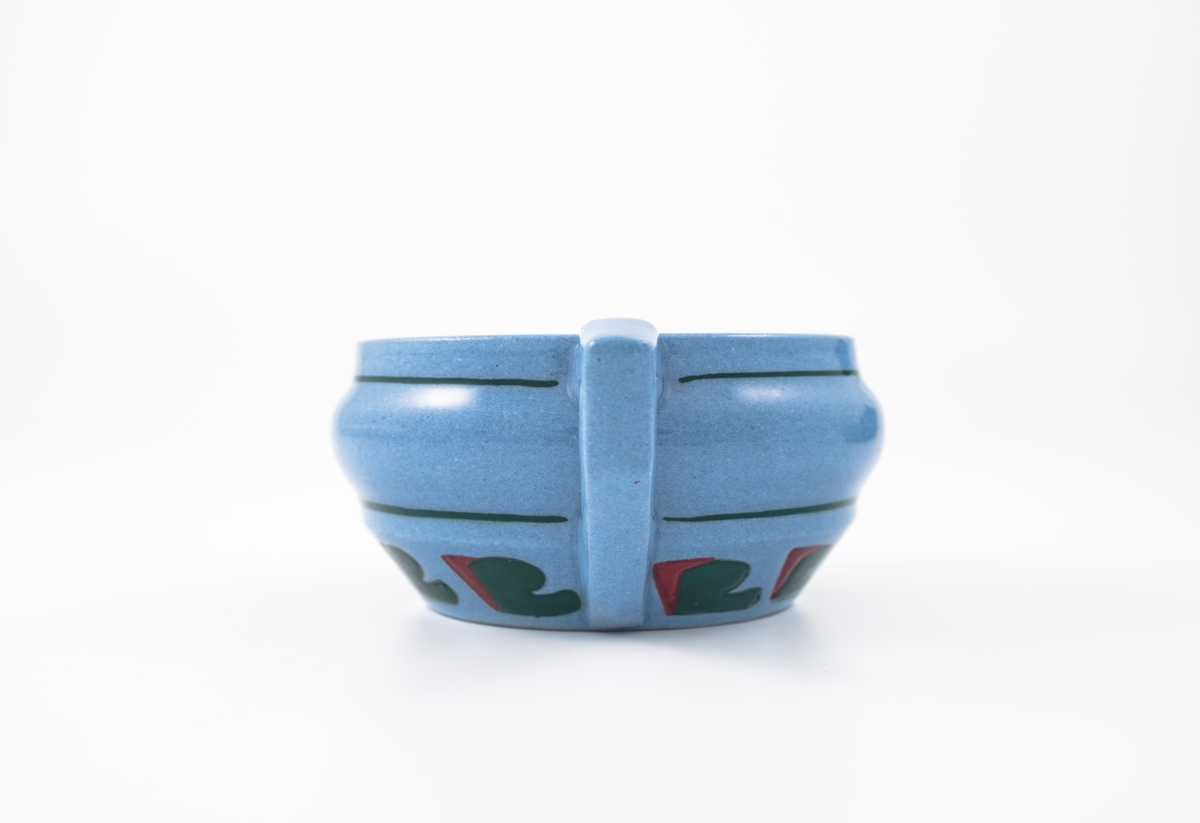 Tekopp med underskål i glasert steingods med begitning. Flattrykket kuleformet korpus, påsatt hank med knekk, samt sirkulær underskål. Kopp og skål er lyseblå og dekorert med en stilisert bord i grønt og rødt, samt horisontale grønne linjer som understreker deres form.